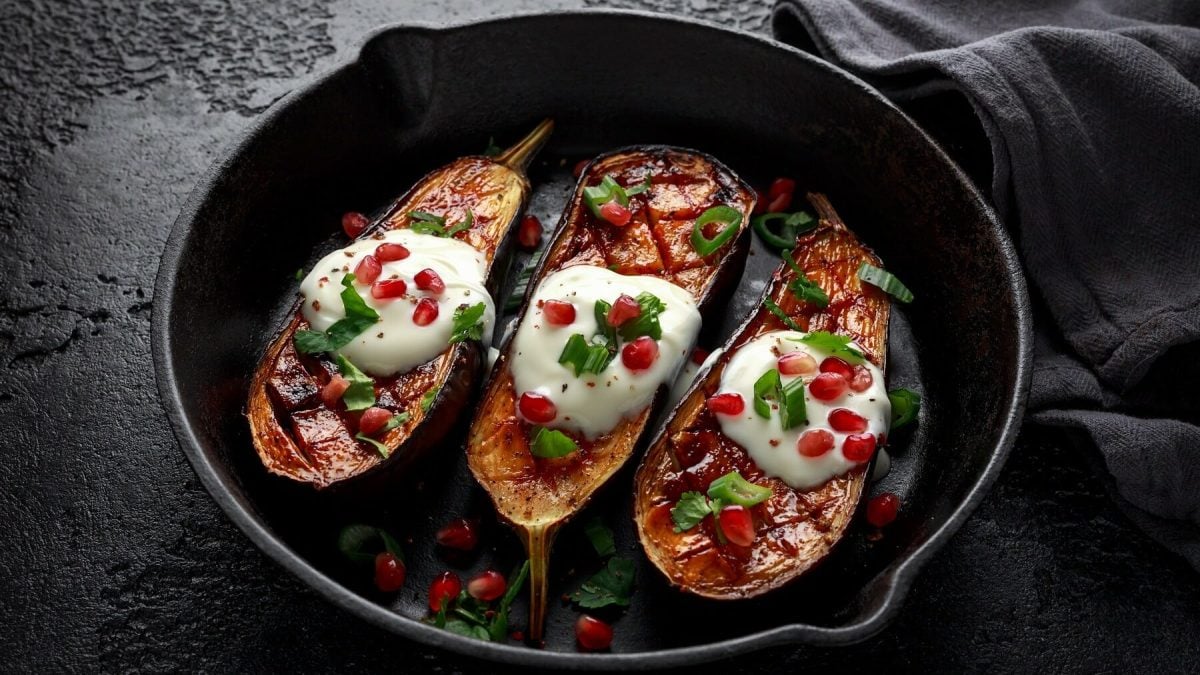 Gebackene Augerginen in einer tiefen schwarzen Pfanne mit Joghurt, Granatapfel und Lauchzwiebeln auf einem schwarzen Tisch neben einem schwarzen Tischtuch.