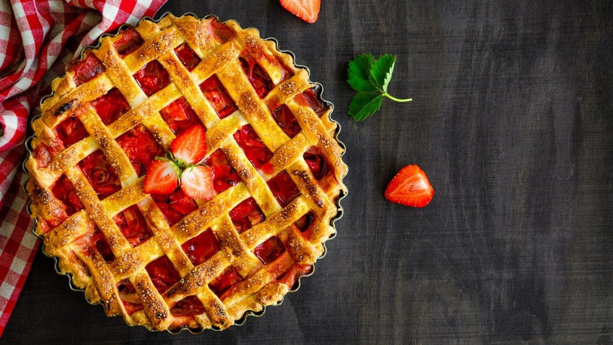 Gitterkuchen mit Erdbeeren und Rhabarber in einer Kuchenform auf einem schwarzen Holztisch neben einer karierten Tischdecke.