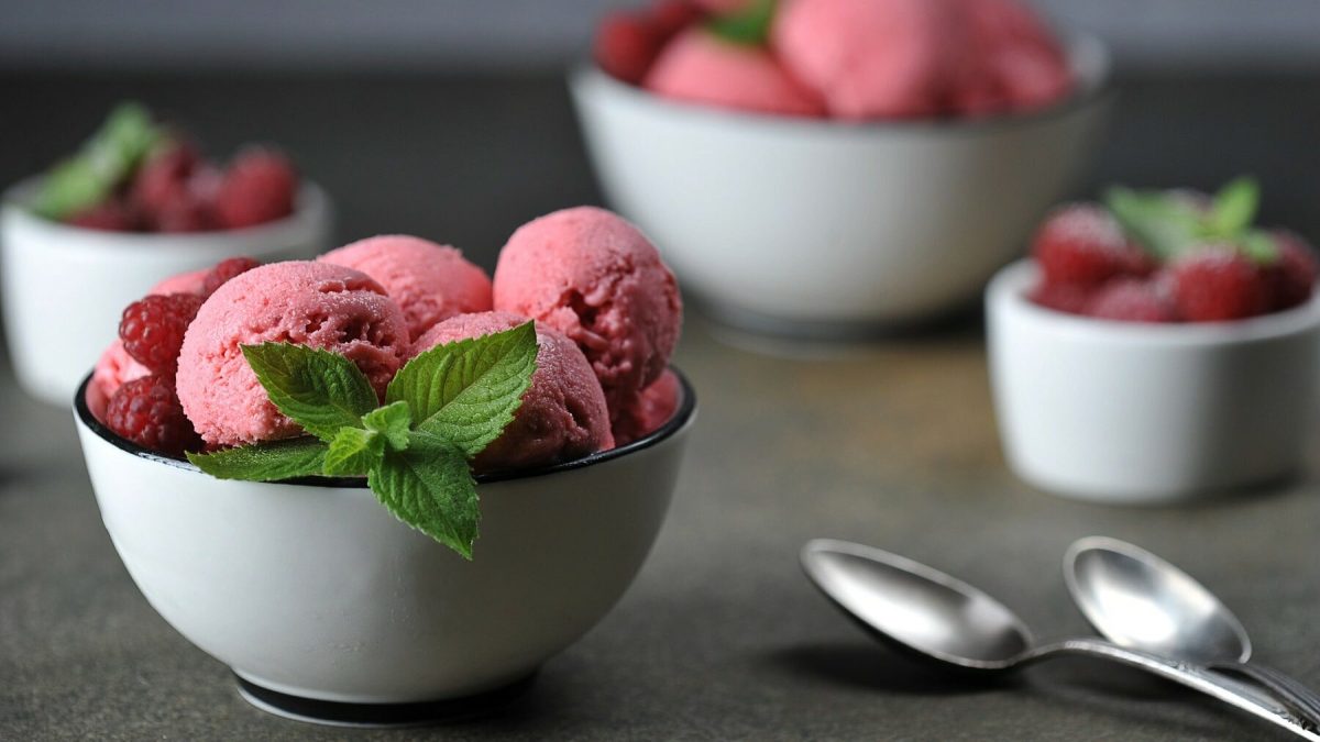 Mehrere Schüsseln mit Himbeer-Joghurt-Sorbet stehen auf einem grauen Untergrund. Die Schüsseln sind weiß und das Sorbet ist mit Minzblättern und Himbeeren garniert. In der rechten unteren Bildecke liegen zwei Löffel.