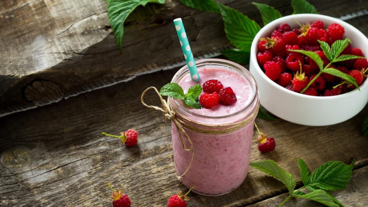 Himbeer-Smoothie mit Banane in einem Glas mit frischer Minze und Himbeeren auf einem Holztisch neben einer Schüssel mit Himbeeren.