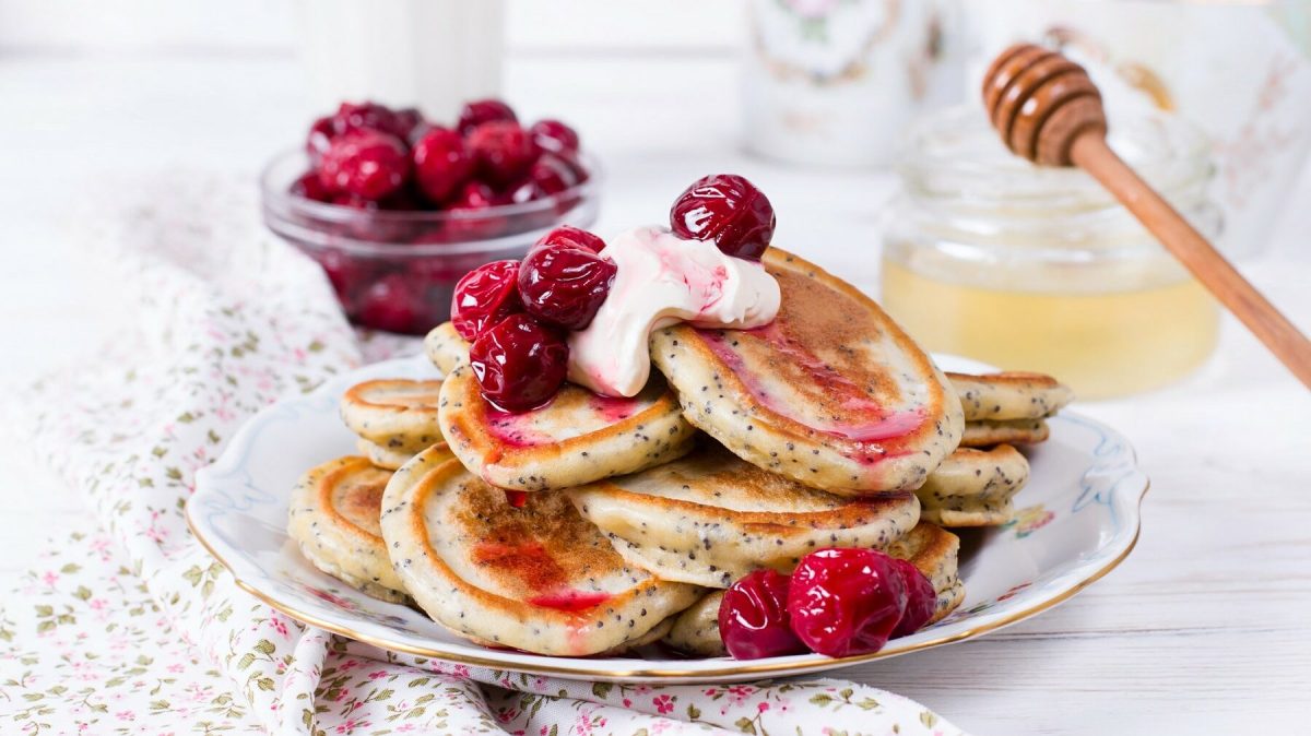 Kirschpfannkuchen mit Mohn und einem Klecks sind serviert auf einem Teller mit Goldrand. Die Pfannkuchen sind mit etwas Sahne angerichtet. Im Hintergrund liegt eine Schüssel mit Sauerkirschen und eine Schüssel mit Honig.