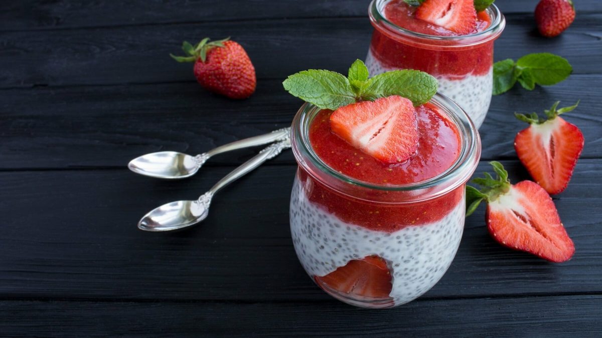 Minzpudding mit Chia und Erdbeeren in kleinen Dessertgläsern neben Löffen auf einem dunklen Tisch.