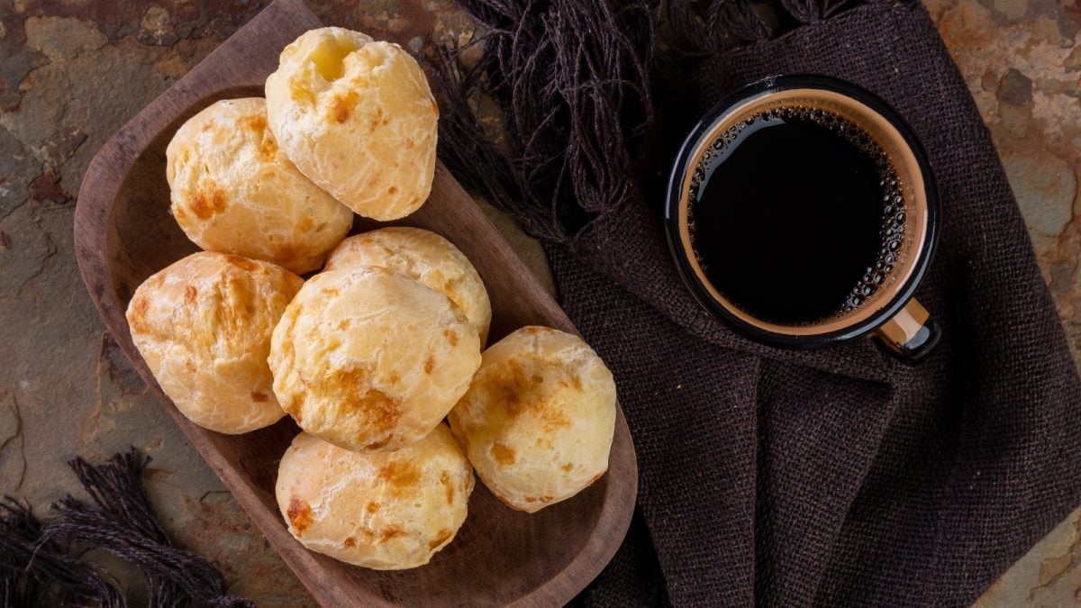 Eine Holzschale mit mehreren Pão de Queijo neben einer Tasse Kafee, von oben, vor dunklem Hintergrund.