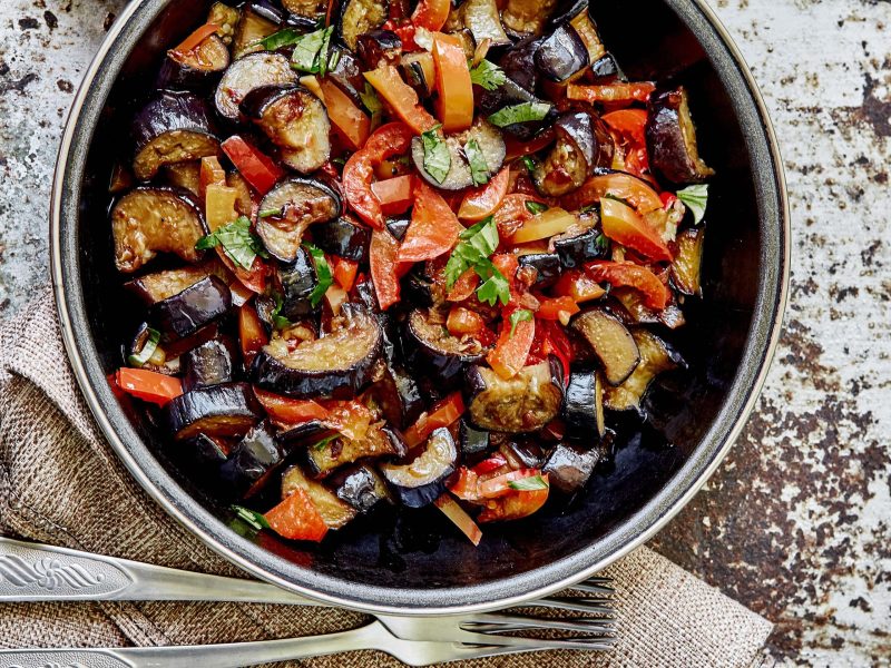 Paprika-Auberginen-Pfanne und einer dunklen Schale. Daneben Besteck.