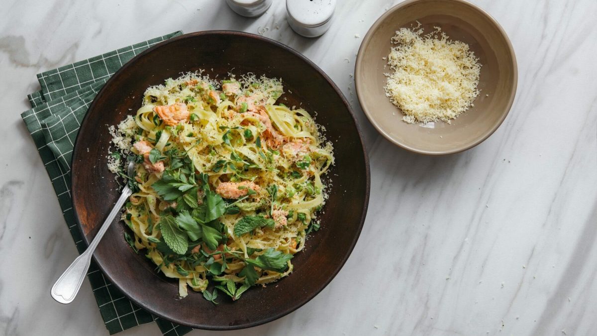 Ein großer, brauner Teller gefüllt mit einer Pasta-Lachs-Pfanne steht auf einem grün karierten Geschirrtuch auf einem Mamortisch. In der Schüssel steckt eine Gabel. Neben der Schüssel steht eine weitere, kleinere Schüssel mit Parmesan und jeweils ein Salz- und Pefferstreuer.
