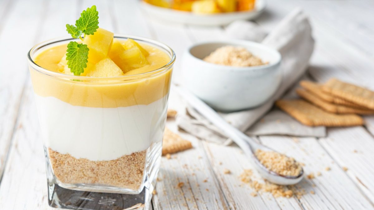 Ricotta-Creme mit Apfelkompott im Dessertglas auf weißem Holz, im Hintergrund Butterkekse, ein Löffel und eine Schale mit Kekskrümeln.