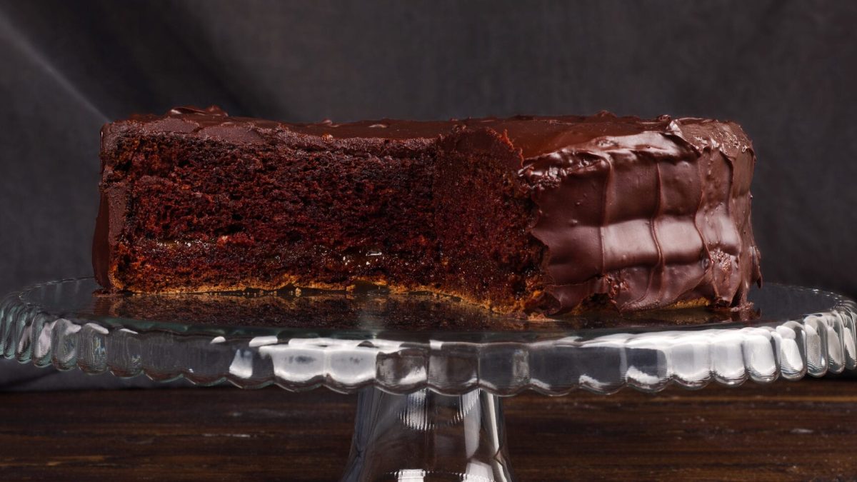 Eine angeschnittene Rum-Schokotorte auf einem Tortenständer aus Glas vor dunklem Hintergrund.