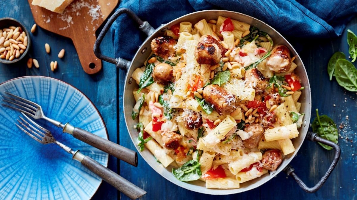 Ein großer Kochtopf mit den Rigatoni mit Salsiccia, Tomaten und Spinat und Pinienkernen neben einem Teller mit Gabel und Parmesankäse, von oben vor blauem Hintergrund.