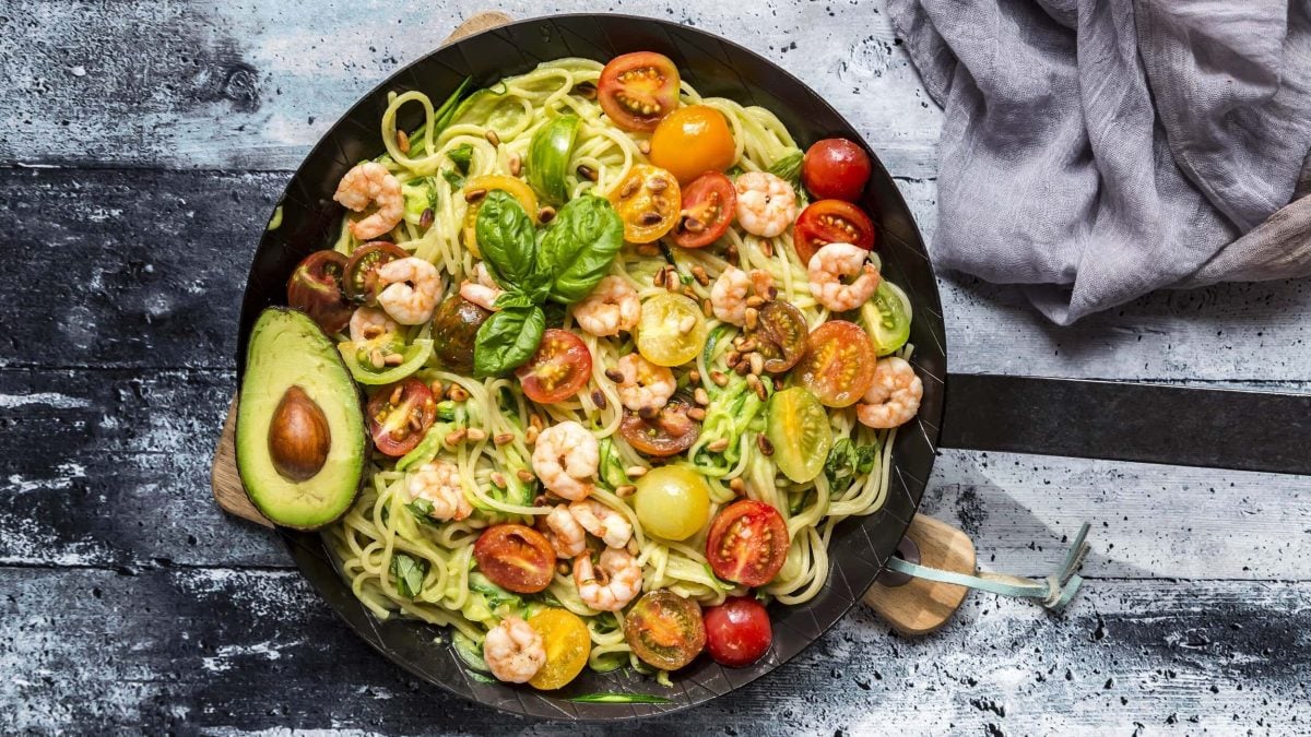 Spaghetti mit Garnelen, Avocado und Tomaten in einer schwarzen Pfanne auf einem gräulichen Holztisch neben einer grauen Tischdecke.