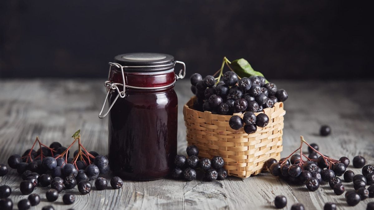 Ein Glas Aroniamarmelade mit Rotwein, daneben steht ein kleines Körbchen mit Aroniabeeren.