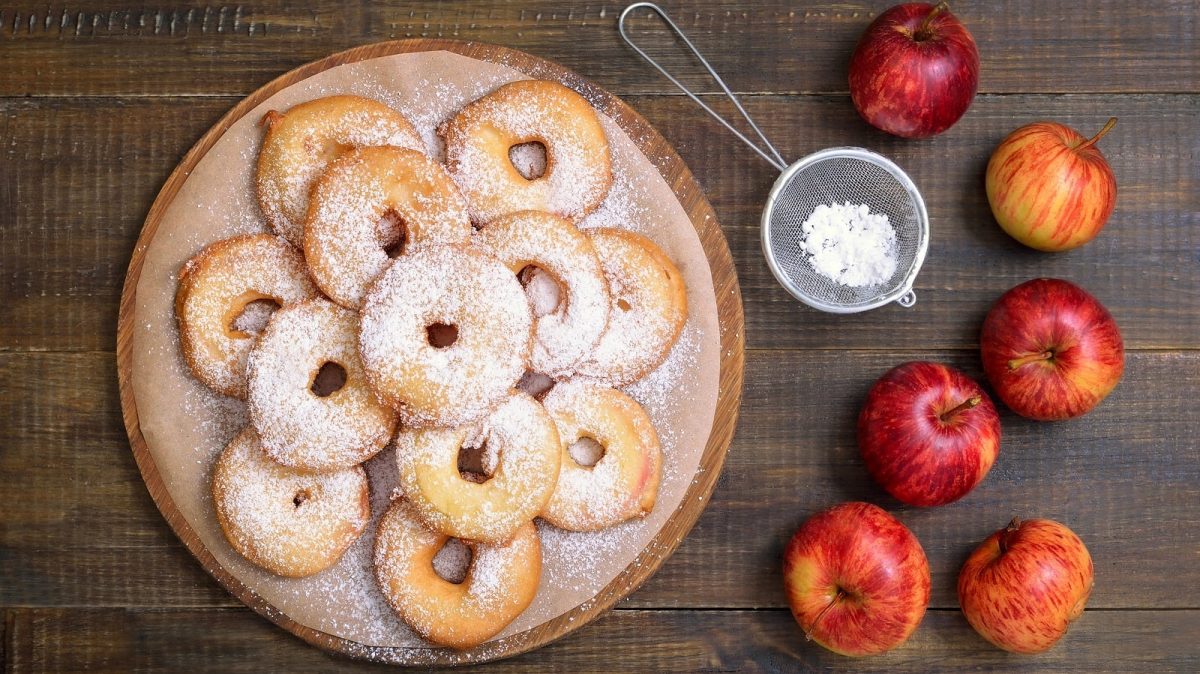 Ein rundes Holzbrett mit aufeinandergestapelten Frittelle di Mele, die mit Puderzucker bestreut sind, neben ganzen Äpfeln und einem kleinen Sieb vor einem Holzhintergrund von oben.