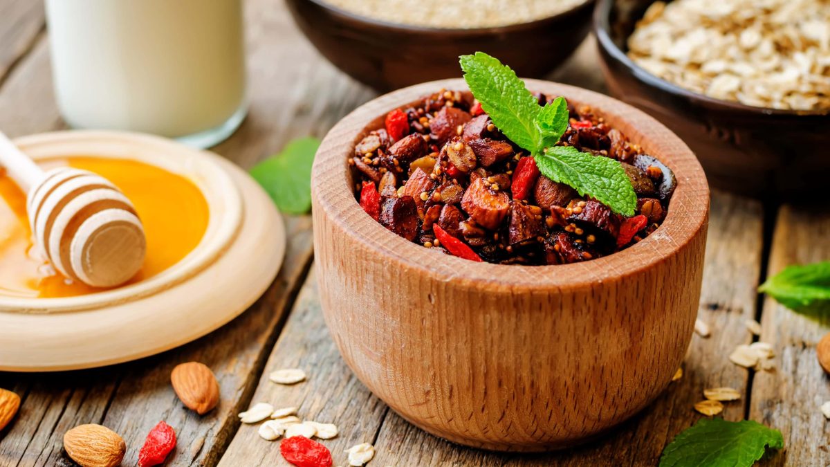 Granola mit Quinoa und Heidelbeeren in einer hellen Holzschüssel neben Honig und Haferflocken auf einem hellen Holztisch.