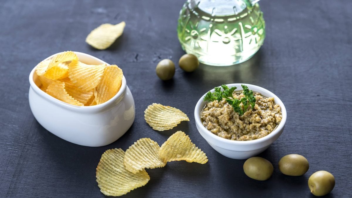 Grüne Oliventapenade in einer kleinen weißen Schüssel auf einem schwarzen Holztisch neben kartoffelchips, frischen Oliven und einer Karaffe mit Weißwein.