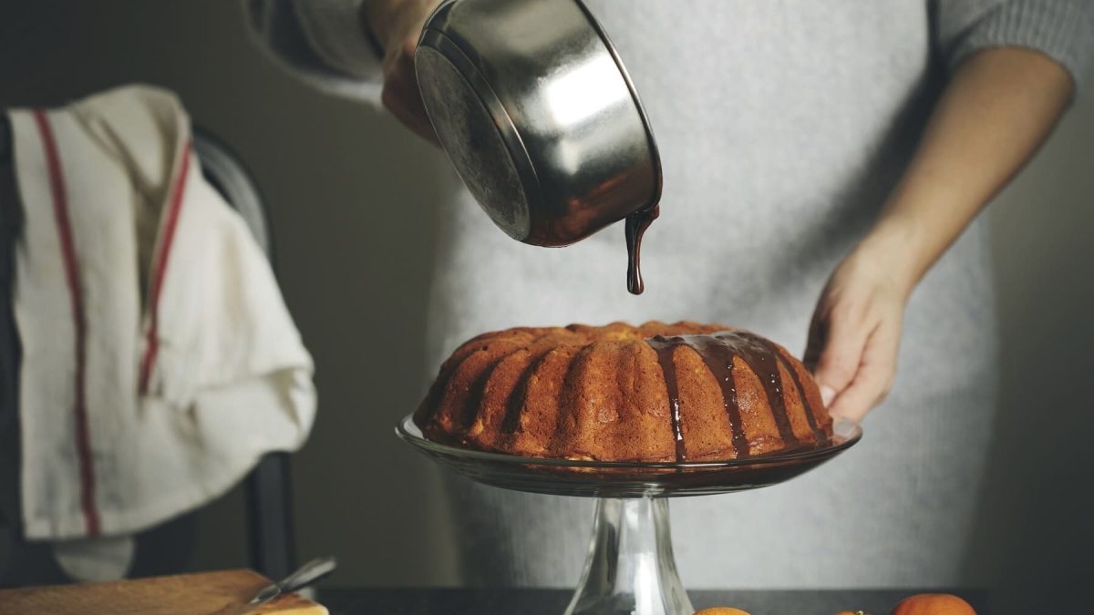 Ein Kuchen steht auf einem Kuchenständer auf einem schwarzen Tisch. Auf dem Tisch liegen Mandarinen und ein Holzbrett. Eine Frau in einem Wollkleid gießt Schokoglasur über den Kuchen.