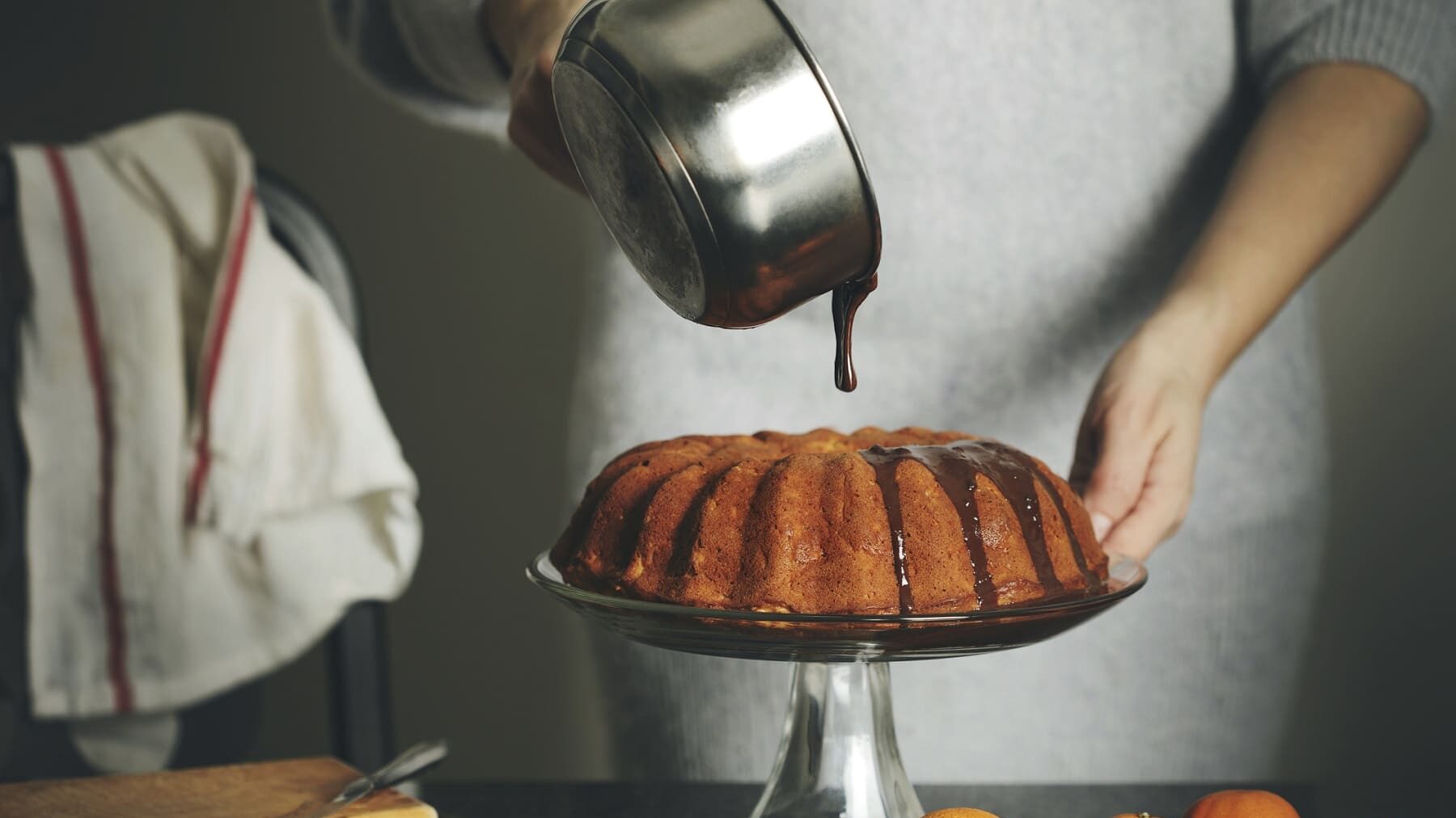 Ein Kuchen steht auf einem Kuchenständer auf einem schwarzen Tisch. Auf dem Tisch liegen Mandarinen und ein Holzbrett. Eine Frau in einem Wollkleid gießt Schokoglasur über den Kuchen.