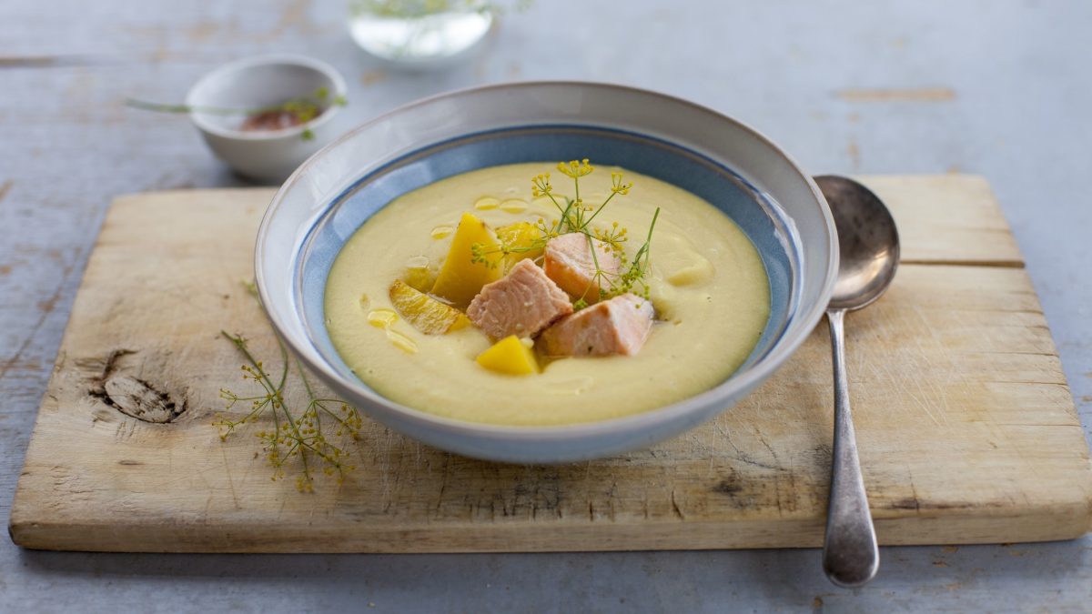 Kartoffelsuppe mit Lachs in einer Schüssel. Daneben ein Löffel. Das Ganze steht auf einem Holzbrett.