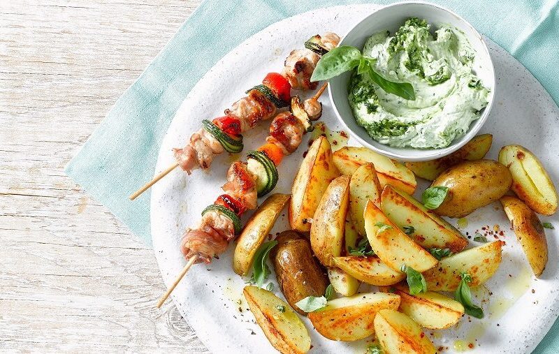 Knusprige Kartoffelecken mit Bärlauch-Dip und Grillspießchen auf weißem Teller vor blauem Tuch.