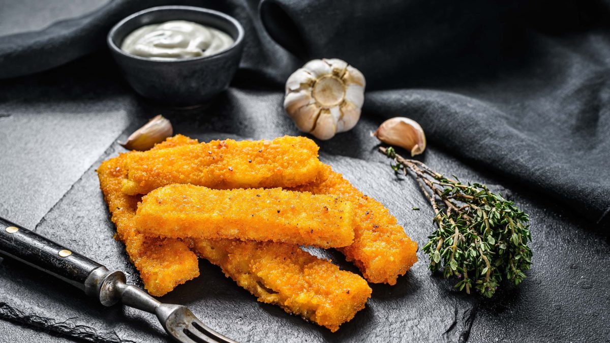Fischstäbchen auf einer schwarzen Schieferplatte neben frischen Thymian, Knoblauch und einer kleinen Schüssel mit einem Knoblauchdip.