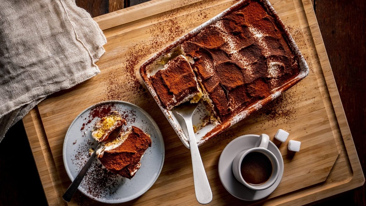 Eine Auflaufform mit klassischem Tiramisu von dem ein Stück herausgeschnitten wurde, das auf dem Teller daneben liegt. Unterhalb der Auflaufform steht eine Tasse Espresso mit zwei Würfeln Zucker. Alles steht auf einem Holzbrett, von oben fotografiert.