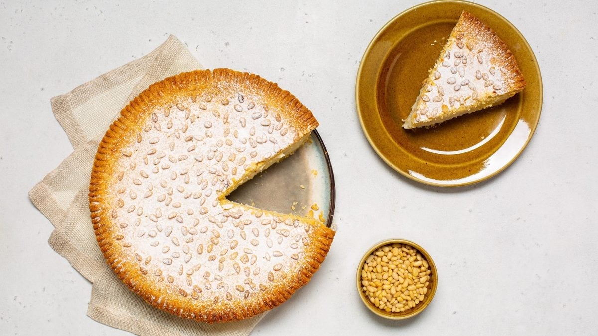 Die runde Torta della Nonna auf einem Teller mit einem Stück abgeschnitten, das auf einem weiteren Teller daneben steht, sowie eine kleine Schüssel mit Pinienkernen, von oben, vor hellem Hintergrund.