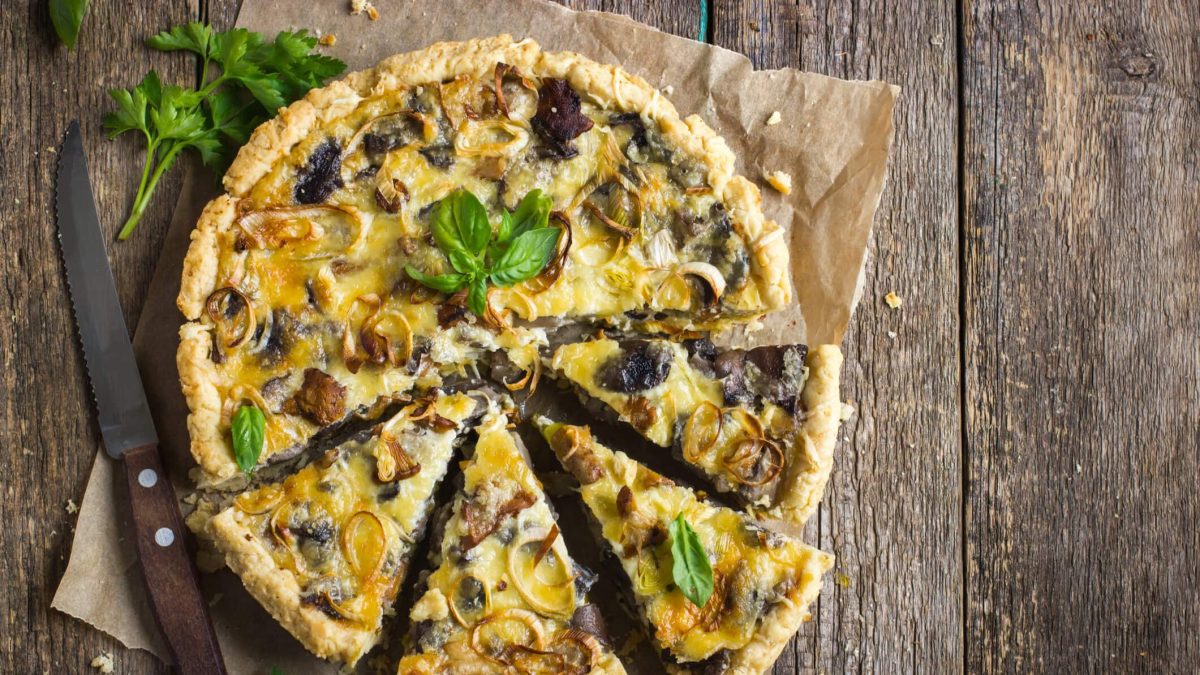 Eine vegetarische Pilzquiche mit Lauch, Käse und Basilikum auf einem Stück Backpapier auf einem rustikalen Untergrund, daneben ein Messer, etwas Knoblauch und Petersilie.