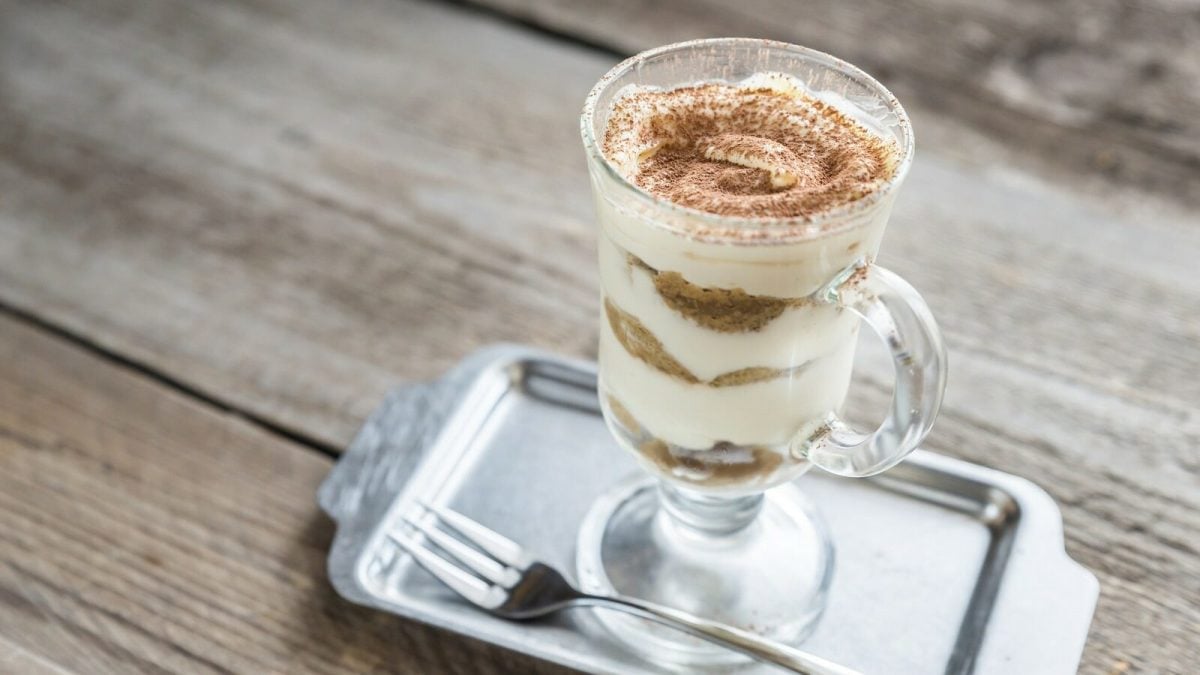 Eine Portion Bananen-Tiramisu un einem Glas steht auf einem silbernen Tablett auf einem braunen Holztisch. Neben dem Glas liegt eine silberne Gabel. Das Tiramisu ist angerichtet mit Kakaopulver.