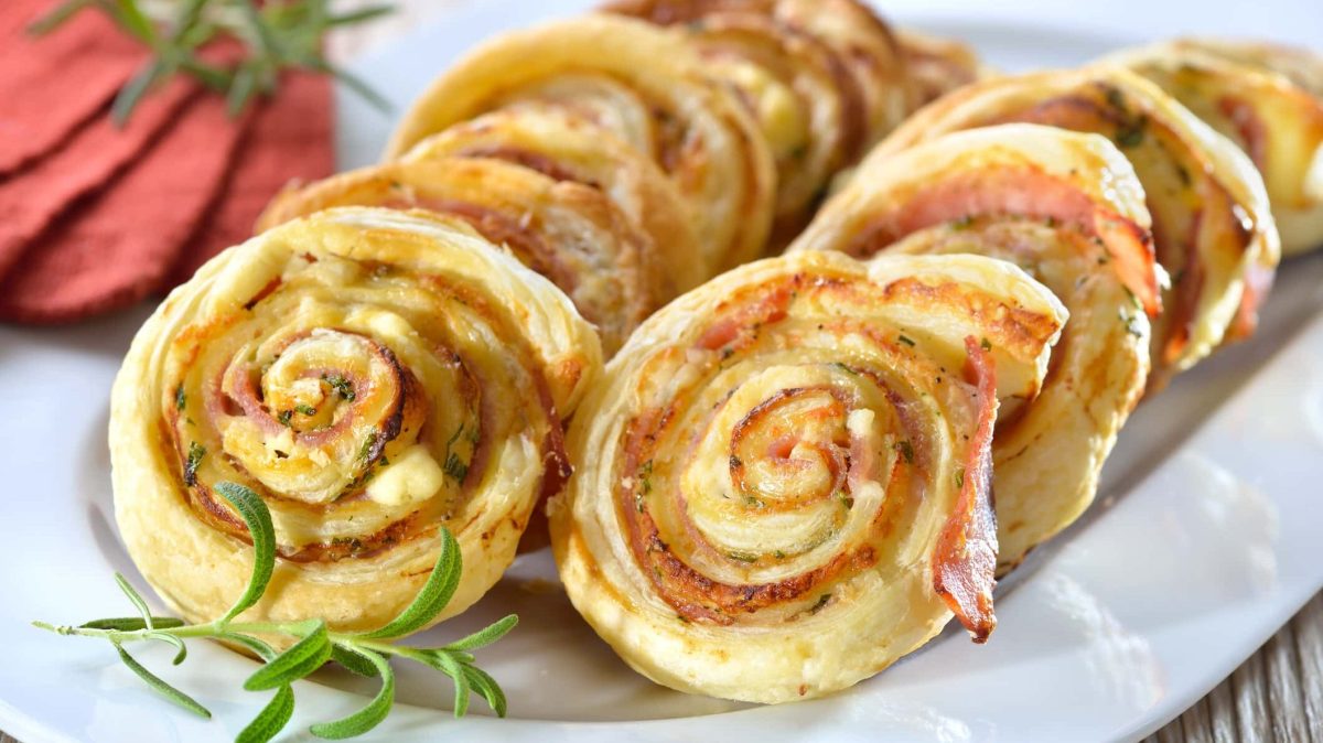 Mit Schinken, Käse und Kräutern gefüllte Blätterteigschnecken auf weißer Servierplatte.