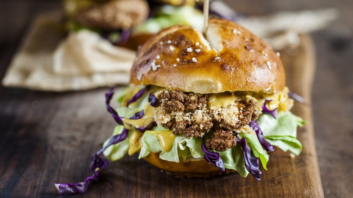 Ein Burger mit Blumenkohl-Patty steht auf einem raunen Holzbrett auf einem braunen Untergrund. Im Hintergrund steht ein weiterer Burger.