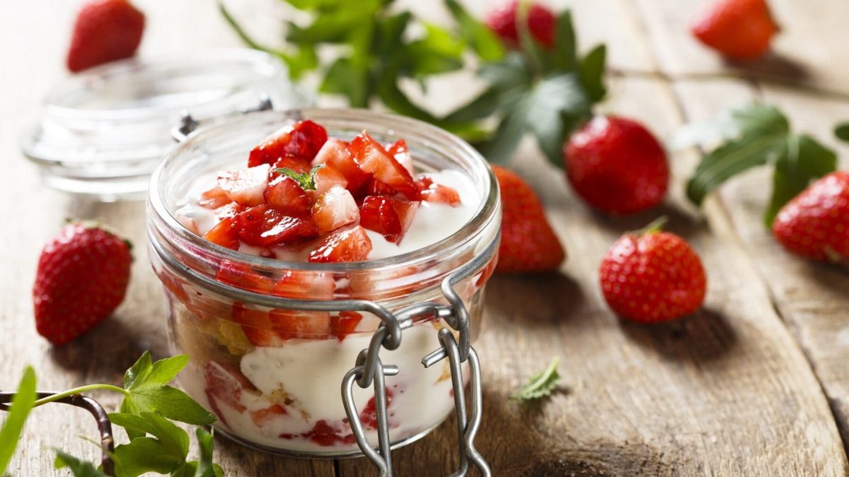Eine Portion Erdbeer-Tiramisu im Glas steht auf einem Holztisch. Das Tiramisu ist angerichtet mit Erdbeerstückchen. Im Hintergrund liegen einzelne Erdbeeren und Grün.
