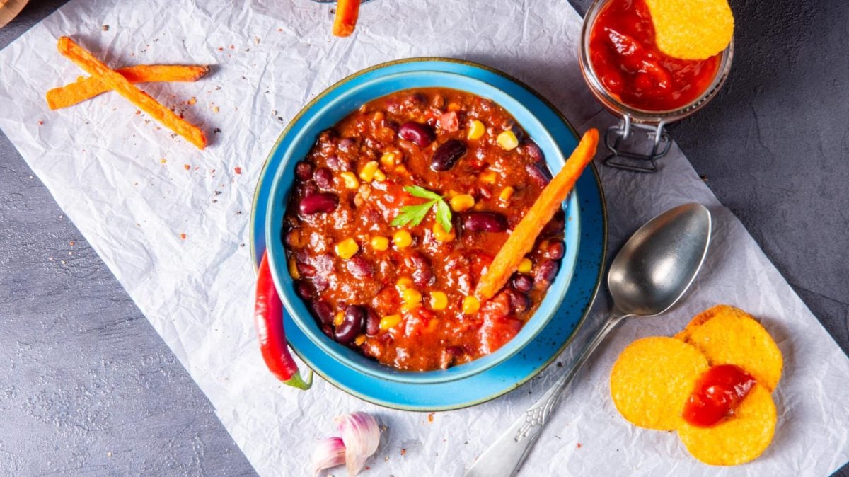 Auf einem rustikalem Tisch liegt ein helles Tuch. Darauf steht eine große Schüssel mit blauem Rand, in der das Süßkartoffel-Chili zu sehen ist. Neben der Schüssel liegen noch Süßkartoffel-Sticks, sowie einige Nachos und ein Glas mit einer roten Soße.