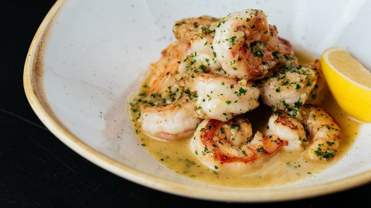 Gambas con Ajo liegen in einer weißen Schüssel mit beigem Rand. Sie sind angerichtet mit Gewürzen und einem Zitronenstück. Der Untergrund ist schwarz.
