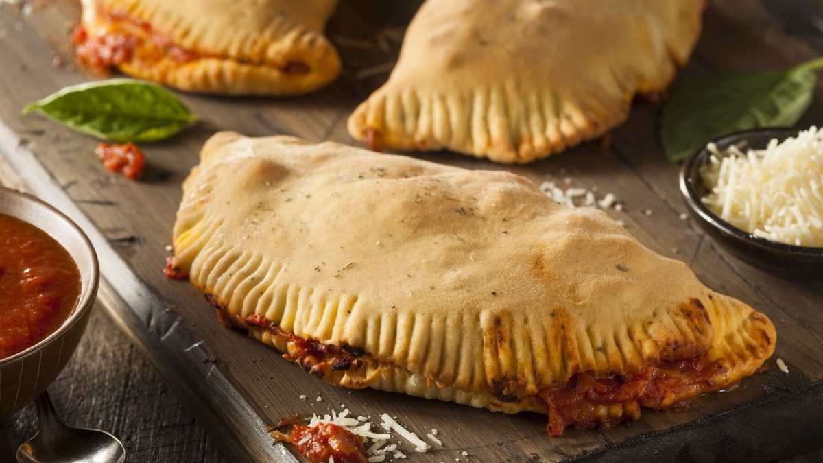3 Gemüse-Schinken-Calzones liegen auf einem Holzbrett. Links neben dem Brett steht ein Gefäß mit Tomatensoße, rechts eine kleine Schale mit Käse.