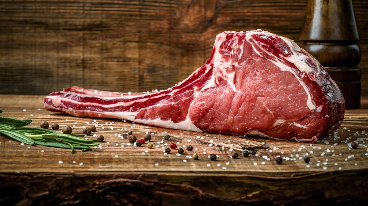 Ein saftig marmoriertes Tomahawk-Steak liegt mit Meersalz und Pfefferkörnern auf einem Holzuntergrund.
