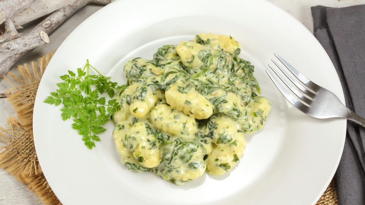 Eine Portion Gnocchi mit Spinat-Béchamel auf einem weißen Teller steht auf einer geflochtenen Matte auf einem Tisch. Am rechten Rand vom Teller liegt eine silberne Gabel.