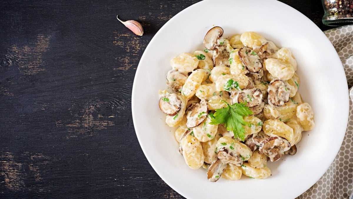 Ein weißer Teller mit Gnocchi con Funghi steht auf einem braunen Holztisch. Neben dem Teller liegt ein gepunktetes Geschirrtuch und eine Knoblauchzehe.