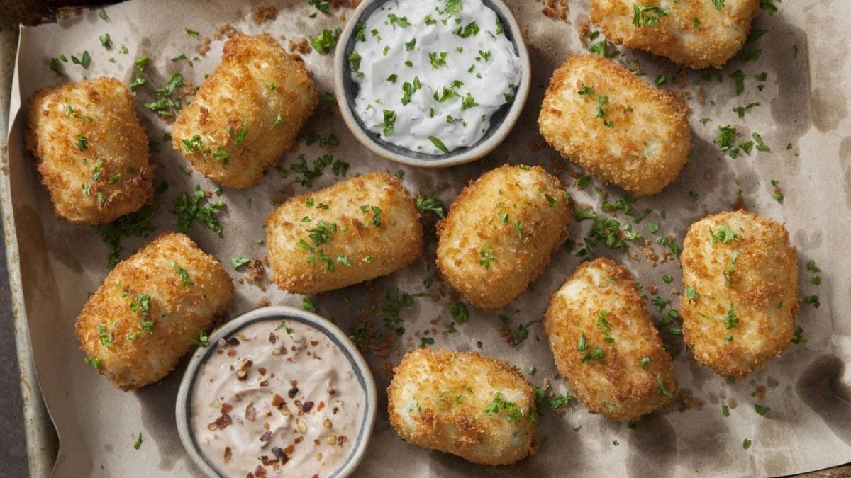 Mehrere Kroketten mit Petersilie liegen auf einem Blech mit Backpapier. Neben den Kroketten stehen verschiedene Dips.