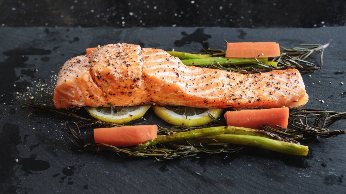 Ein Stück Lachsfilet liegt auf Rosmarin, Zitronenscheiben, Spargel und Karotten. Der Untergrund ist eine schwarze Steinplatte. Über dem Lachs und dem Gemüse sind Salz und Pfefferkörner verteilt.