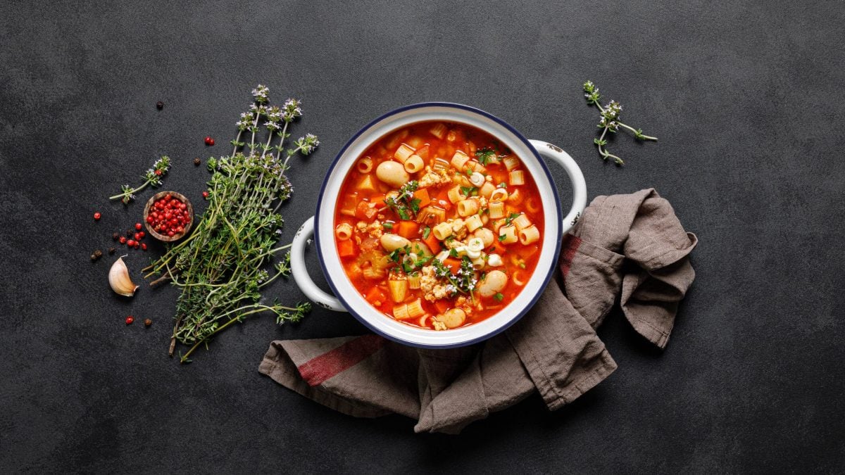 Italienische Minestrone mit Pasta und Bohnen angerichtet auf dunkler Schieferplatte, umgeben von Zutaten. Platte