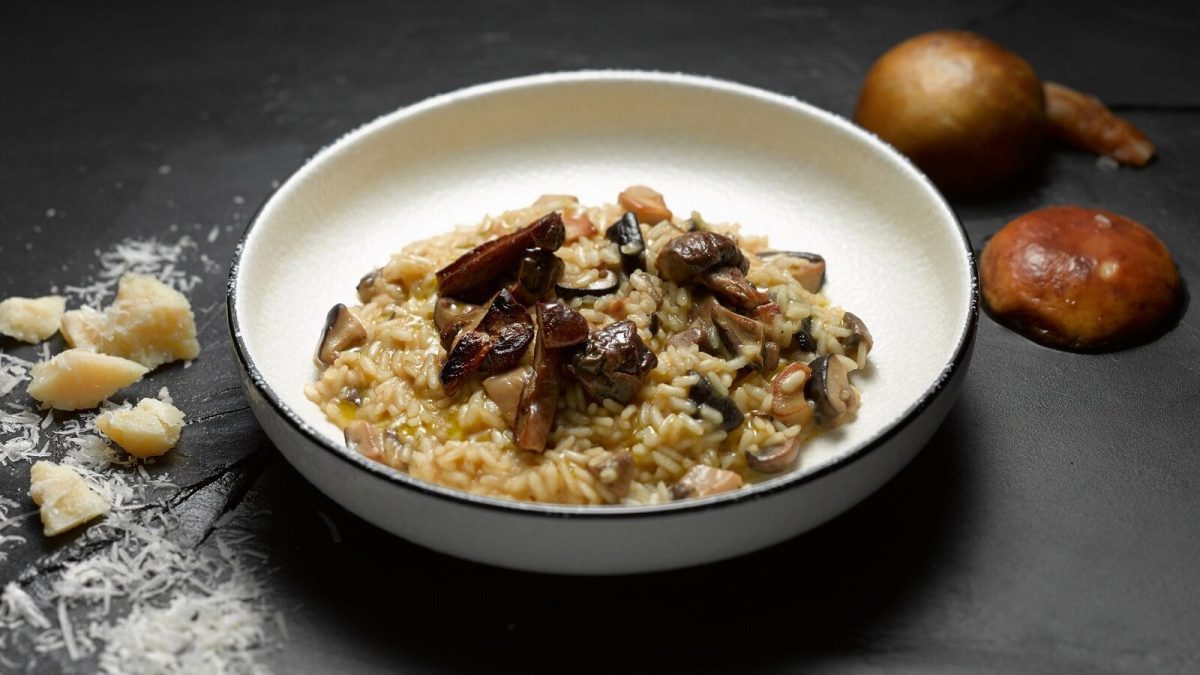 Eine Schale mit Pilzrisotto und Parmesan steht auf grauem Hintergrund. Um die Schale herum liegen Pilze mit Parmesan.