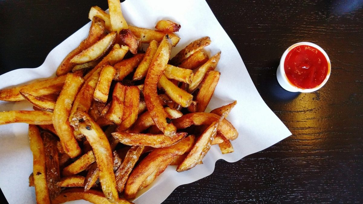 Eine Portion Pommes auf einer weißen Servierte liegt auf einem schwarzen Holztisch, Neben den Pommes steht ein kleiner Becher mit Ketchup.
