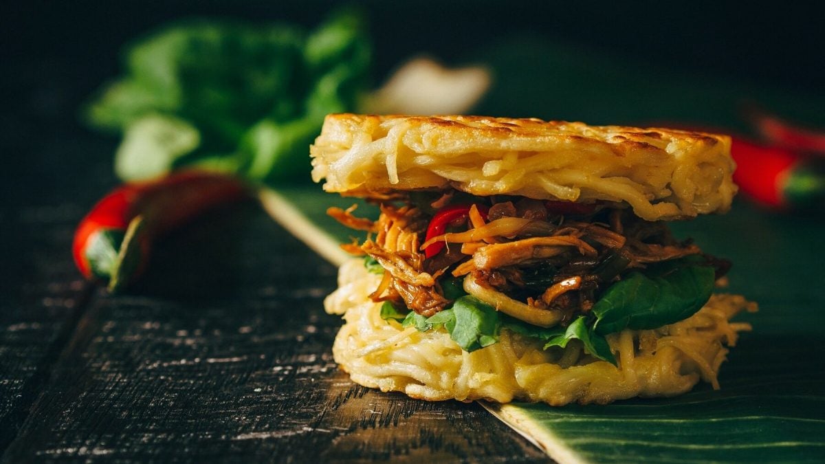 Ein Ramen-Burger steht auf einem Bananenblatt auf einem schwarzen Tisch. Der Ramen-Burger ist belegt mit Tomaten, Rucola, Hähnchen und Zwiebeln. Im Hintergrund liegt Salat und mehrere Chilichoten.