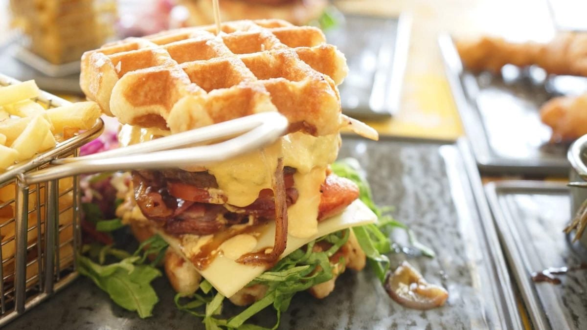 Ein Waffel-Burger liegt auf einer Steinplatte. Der Burger ist belegt mit Zwiebeln, Tomaten, Rucola, Tomaten, einem Burgerpatty und viel Käse. Neben dem Burger steht eine Portion Pommes.