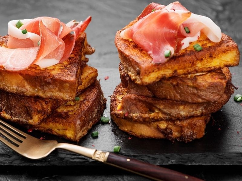 Mehrere Scheiben Armer Ritter liegen auf einer Schieferplatte. Auf den Scheiben liegt Prosciutto. Außerdem befindet sich eine Gabel auf der Platte.