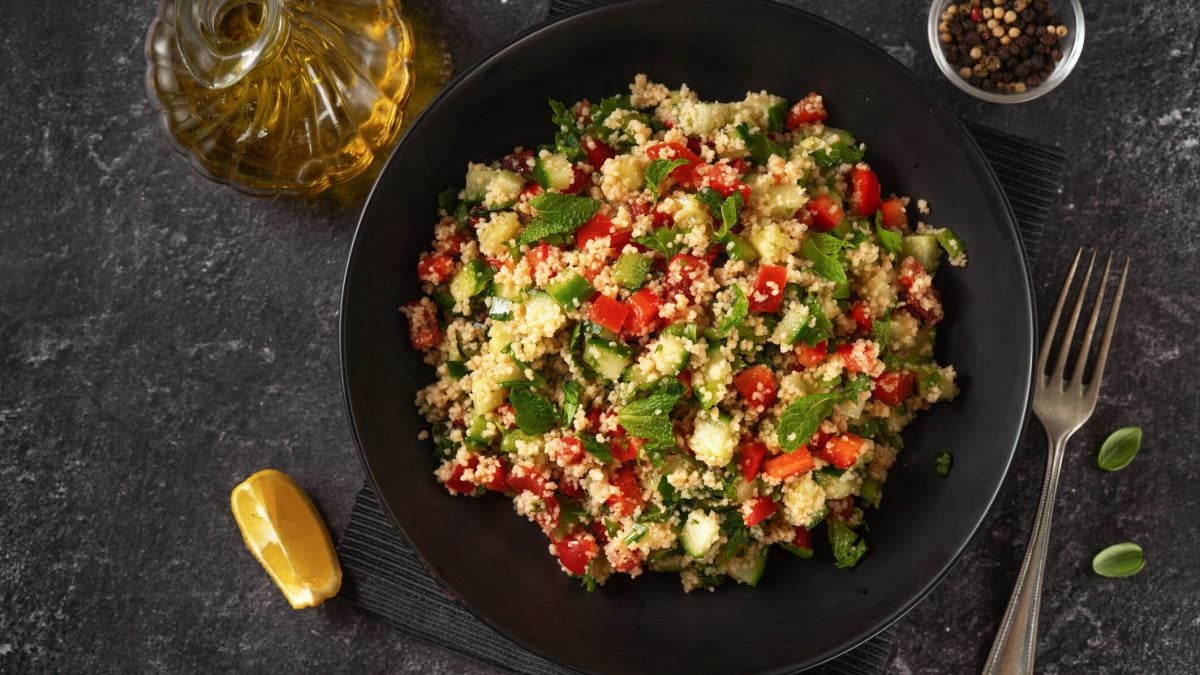 Ein dunklter Teller mit einer Portion Couscous-Tomatensalat, daneben Besteck, eine Zitronenspalte, eine Flasche mit Öl und eine Schale mit Pfefferkörnern.