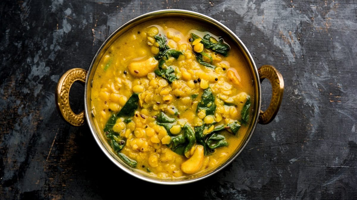 Eine gusseiserne Schüssel mit verzierten Henckeln gefüllt mit gelben Dal Palak, Linsen-Curry mit Spinat, hier verfeinert mit Cashew-Nüssen und Kreuzkümmel, vor einem dunklen Hintergrund von oben.