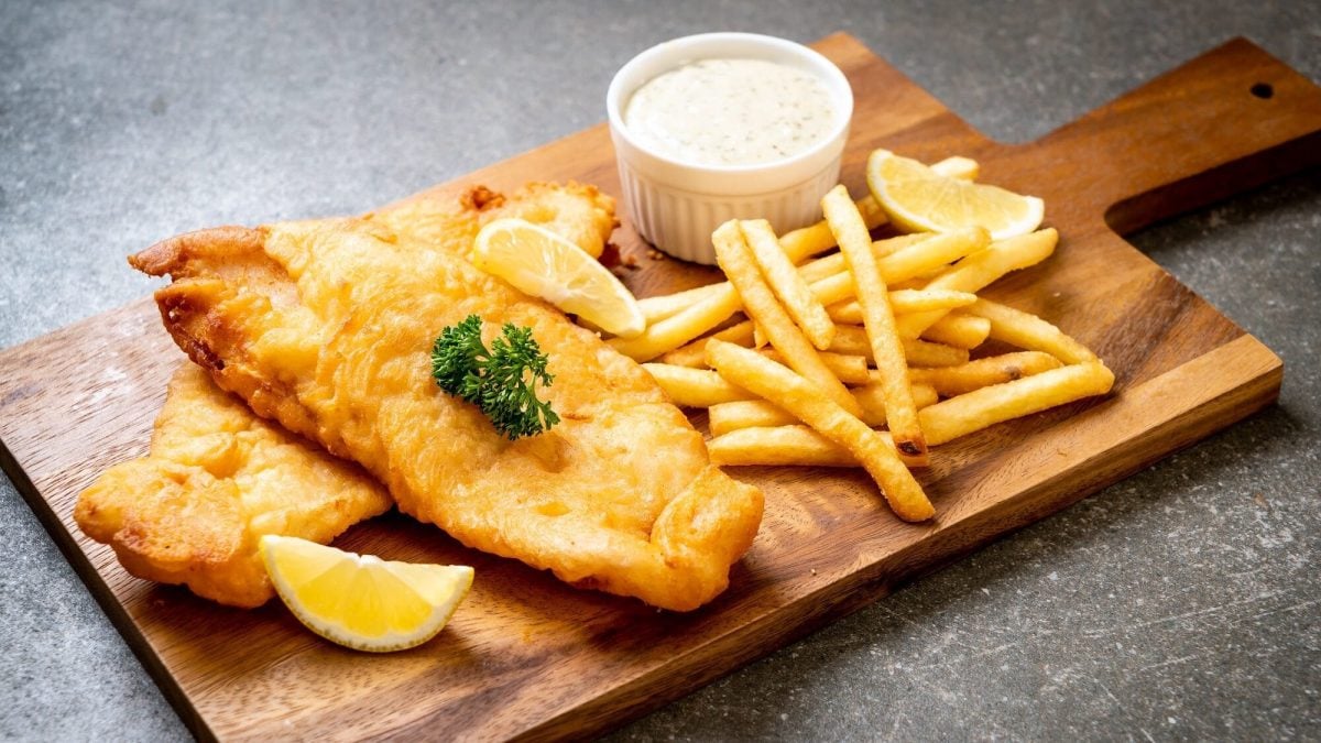 Eine Portion Fish and Chips mit Dip auf Holzbrett, angerichtet auf dunklem Untergrund