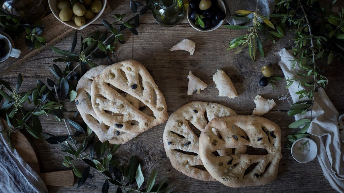 Vier Laib des französischen Brotes Fougasse, deren Form an Blätter erinnert, nebeneinander und aufeinander gestapelt, umrandet von Blättern, Tüchern und Oliven, vor einem dunklen Holzhintergrund, von oben.