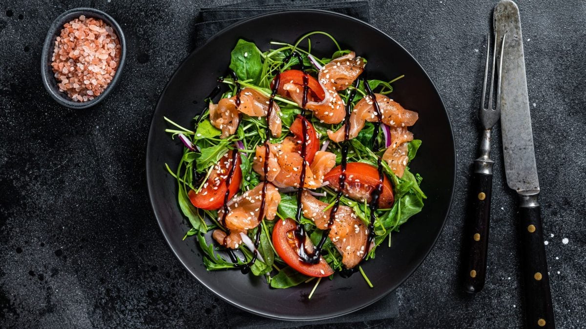 Ein dunkler Teller mit grünem Salat mit Lachs und Tomaten. Daneben liegt Besteck, auf der anderen Seite steht eine Schale mit grobem Salz.