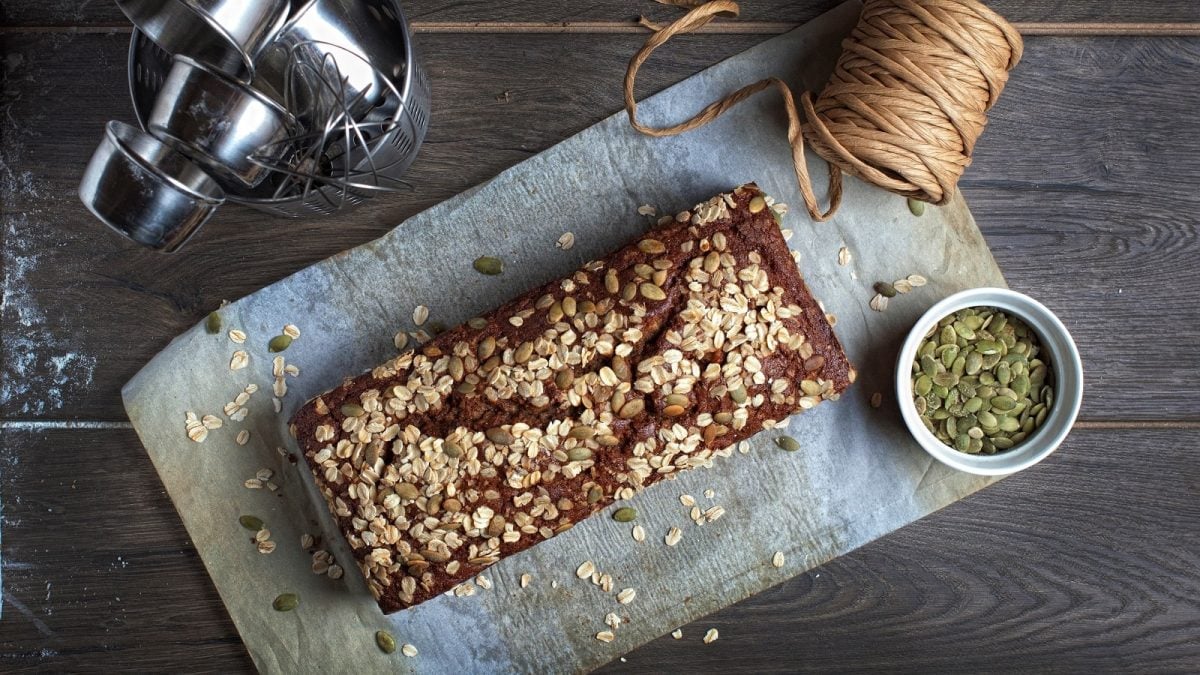 Ein Laib Hanseatenbrot mit Haferflockne und Kürbiskernen bestreut auf einem Backpapier neben einer Schüssel mit Kürbiskernen, einer Rolle mit dicker Schnur und verschiedenen Backutensilien, von oben vor einem Holzhintergrund.