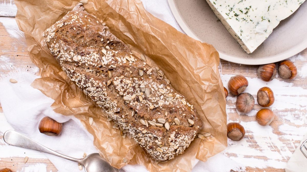 Ein Laib Haselnussbrot mit Haferflocken und Kürbiskernen bestreut auf einem Stück Backpapier neben Haselnüssen und Käse von oben vor einem hellen Holz Hintergrund.